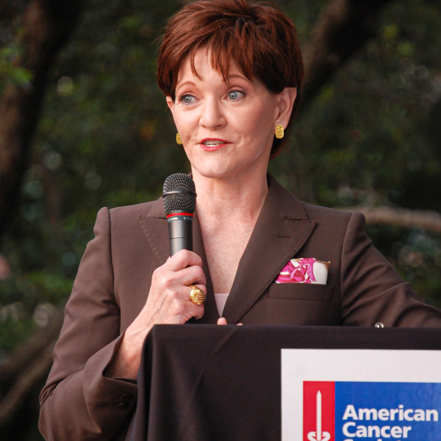 Beth Sanders Moore in a brown suit holding a mic and speaking from a podium at the American Cancer Society Tickled Pink event at Discovery Green.  