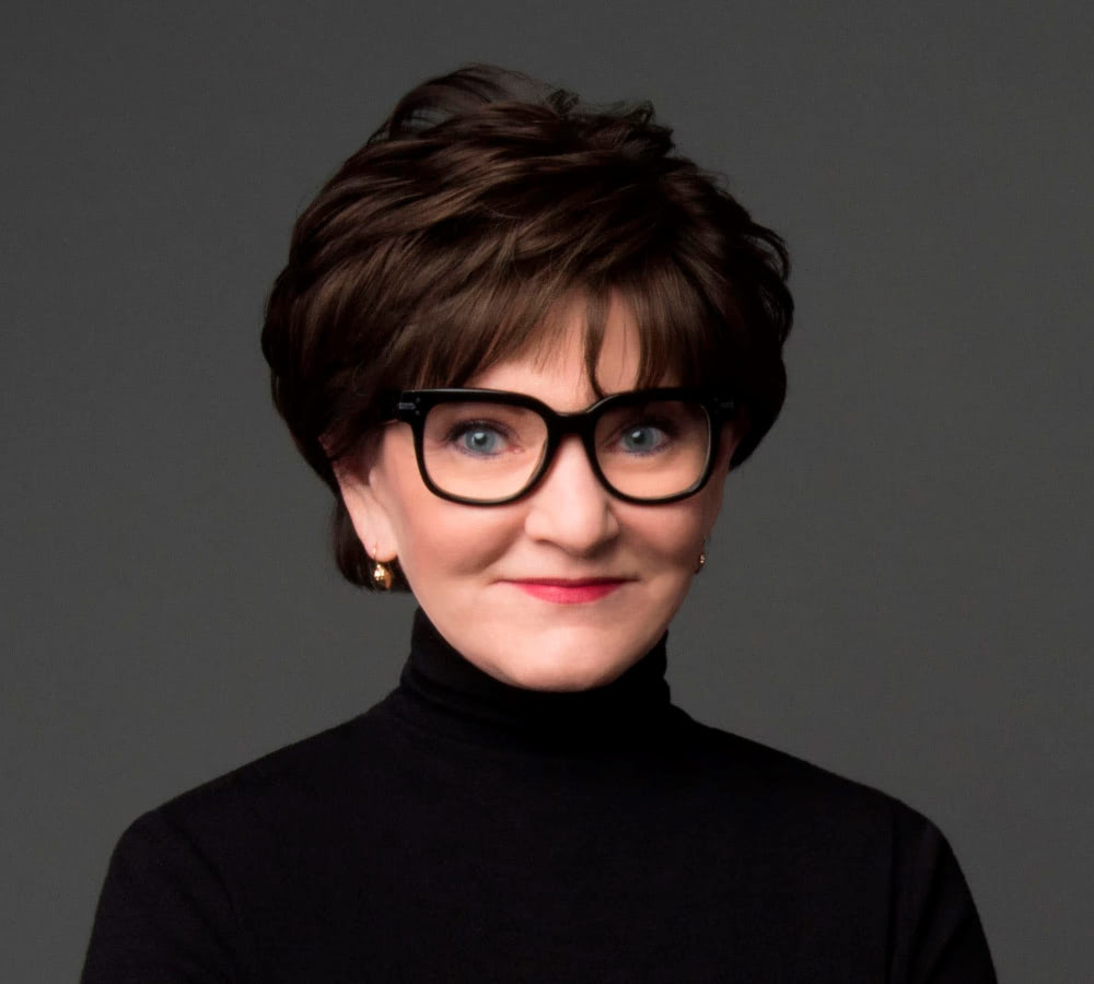 Beth Sanders Moore wearing a black turtleneck sweater and black thick-rimmed glasses posing for a biography headshot.