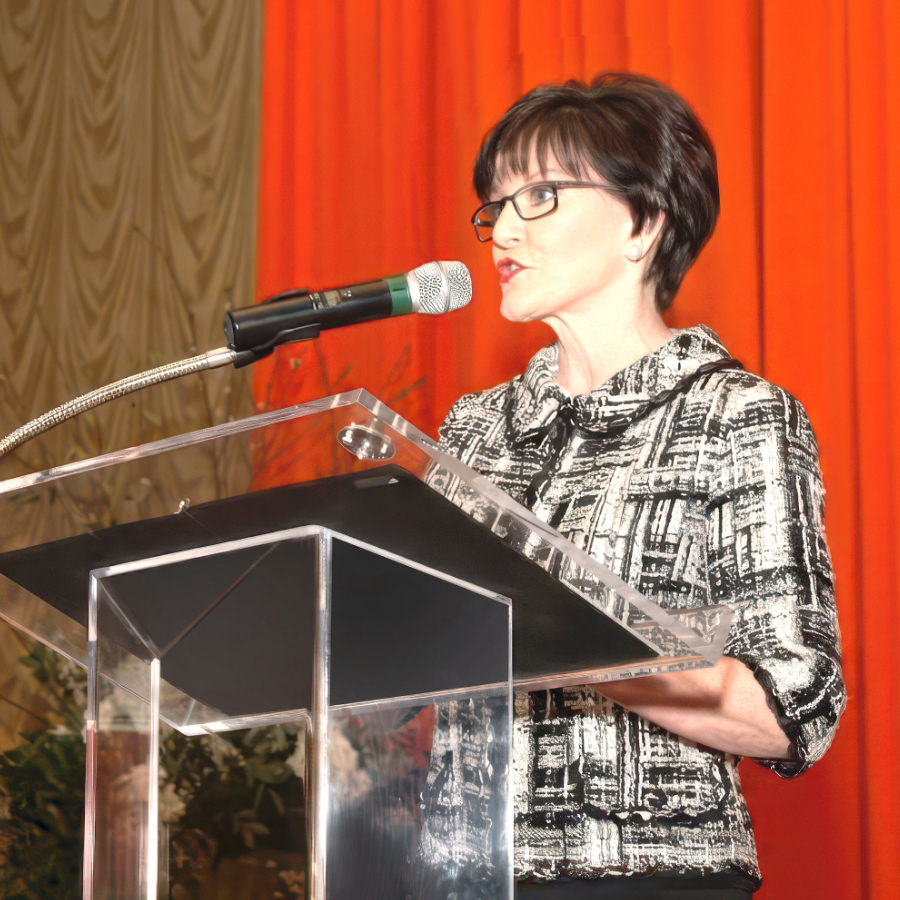 Beth Sanders Moore wearing glasses and tweed jacket standing on stage as moderator of a healthy cancer survivorship forum.