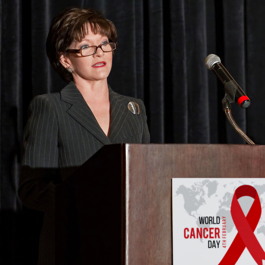 Beth Sanders Moore wearing glasses and pinstripe suit speaking to cancer survivors from a podium displaying the World Cancer Day logo.